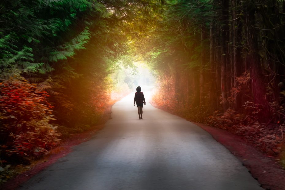 A woman walking on a road toward daylight through a multicolored forest.