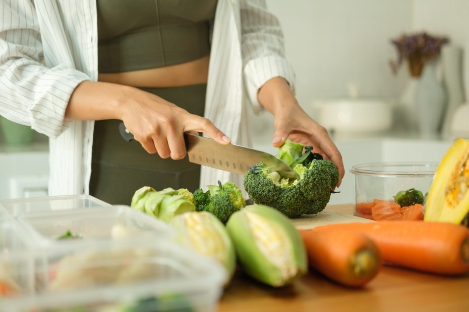 A woman is shown cutting broccoli at a kitchen counter with carrots and cabbage in the foreground
